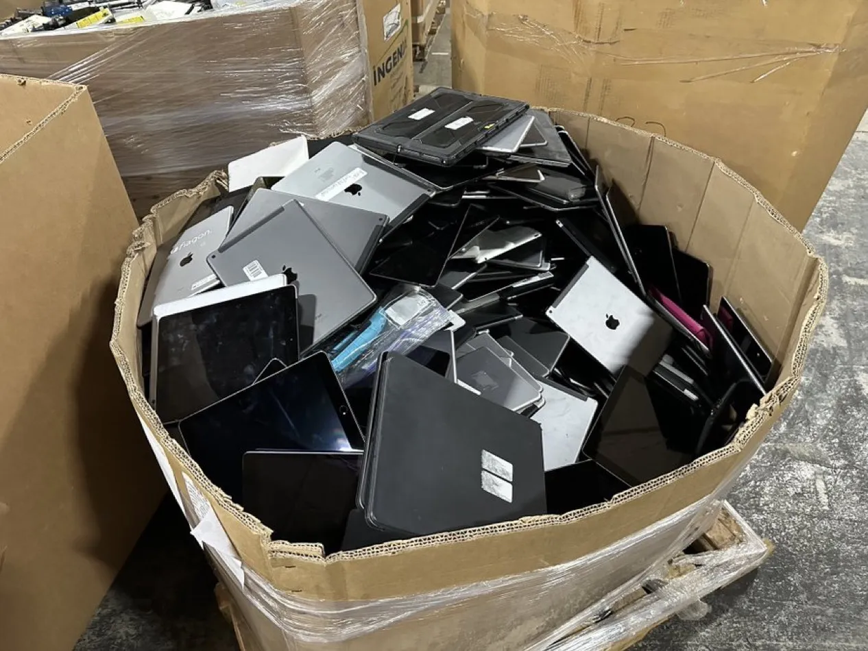 A large cardboard recycling bin overflowing with discarded laptops, iPads, and tablets, waiting to be shredded